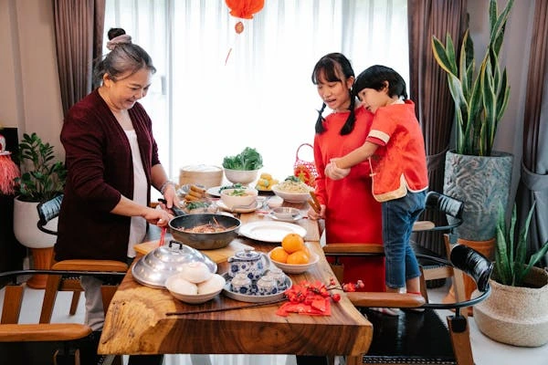 gambar keluarga tionghoa yang sedang berkumpul bersama di meja makan. Mengenakan pakaian serba merah dan sedang mempersiapkan makanan.