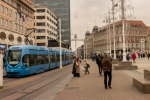 Gambar kota di Jerman, ada kereta listrik warna biru, gedung dan penduduk yang berjalan kaki