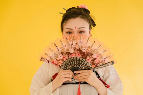 Photo seorang wanita dalam balutan hanfu ala Tiongkok sambil memegang sebuah kipas menutupi setengah wajahnya